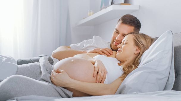 Happy Young Couple Cuddling Together in the Bed, Young Woman is Pregnant and Loving Husband Touches and Caresses Her Belly Tenderly. Camera Focus on the Belly. - Photo, image