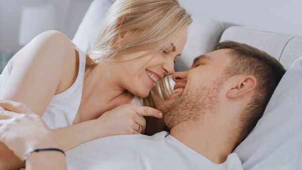 Happy Young Couple Cuddling Together in the Bed, Kissing and Smiling. Beautiful Blonde Female and Her Fit Loving Partner Experiencing Tender and Loving Moments of Living Together. - 写真・画像