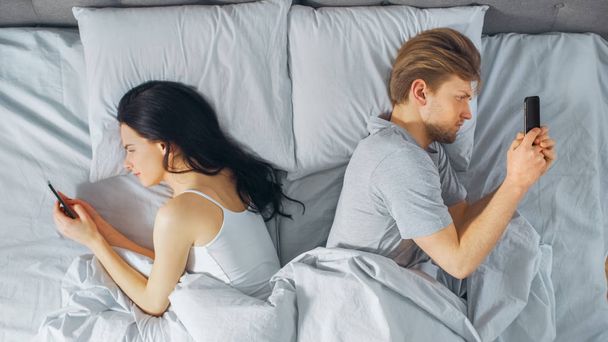 Alienated Millennial Young Couple in the Bed, Young People Turn Away From Each other Using Smartphones, Browsing Through Social Networks and Not Talking to Each Other. Top Down Shot. - Photo, image