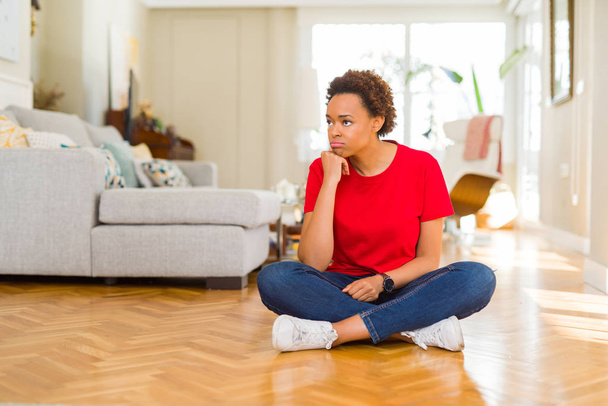 Joven mujer afroamericana hermosa sentada en el suelo en casa pensando que se ve cansada y aburrida con problemas de depresión con los brazos cruzados
. - Foto, imagen