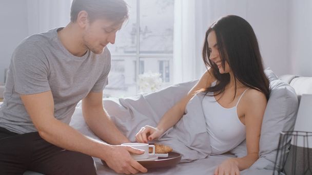 Shot of a Caring Young Man Bringing Tray with Breakfast in Bed to His Lovely Female Partner. Ideal Tender Relationship and Coffee and Croissants in Bed. - Photo, image