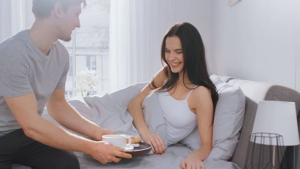 Shot of a Caring Young Man Bringing Tray with Breakfast in Bed to His Lovely Female Partner. Ideal Tender Relationship and Coffee and Croissants in Bed. - Фото, изображение