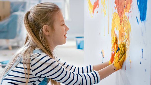 Menina feliz com as mãos mergulhadas na pintura colorida desenha um coração vívido na parede. Ela está se divertindo e rindo. Casa está sendo renovada
. - Foto, Imagem