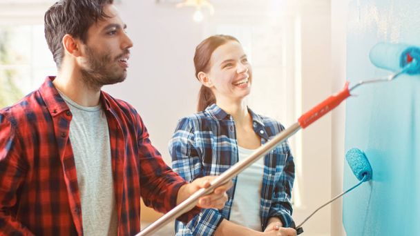 Young Beautiful Couple Decorate Their New Apartment. Husband and Wife are Painting the Wall with Rollers that are Dipped in Light Blue Paint. Renovations at Home. Sunny Ambient Atmosphere. - Foto, Bild