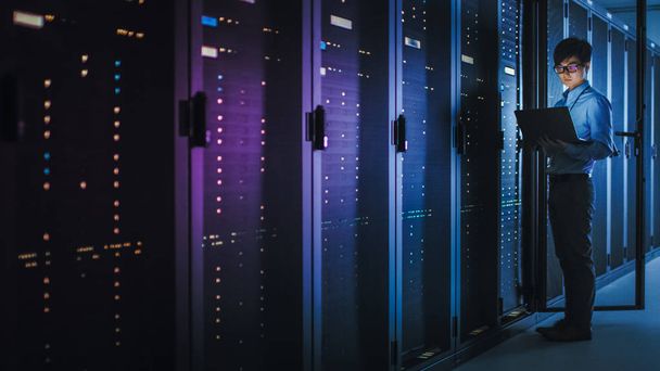 ダークデータセンターで:男性ITスペシャリストは、運用サーバーラックの列の横に立って、メンテナンスのためにラップトップを使用しています。クラウドコンピューティング、人工知能、スーパーコンピュータ、サイバーセキュリティのコンセプトネオン - 写真・画像