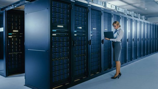 In Data Center: Female IT Technician Stands Near Rack Cabinet and Runs Maintenance Program on Laptop, Controls Operational Server's Optimal Functioning. - Photo, Image