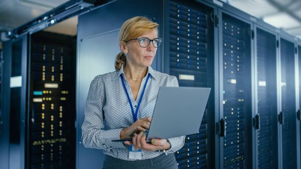 In Data Center: vrouwelijke IT-technicus met onderhoudsprogramma op een laptop, regelt de operationele server rack optimale werking. Modern high-tech telecommunicatie operationeel Data Center. - Foto, afbeelding