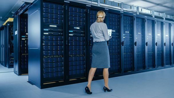 In Data Center: vrouwelijke IT-specialist loopt langs de rij van operationele server racks, gebruikt laptop om het onderhoudsprogramma uit te voeren. Modern high-tech telecommunicatie operationeel Data Center. - Foto, afbeelding