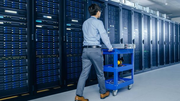 In the Modern Data Center: IT Technician Working with Server Racks, Pushes Cart Between Rows of Server Racks. On a Pushcart New Hardware for System Update. Engineer Doing Maintenance and Diagnostics. - Photo, Image