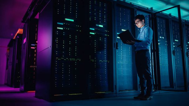 In Data Center: Male IT Technician Running Maintenance Programme on a Laptop, Controls Operational Server Rack Optimal Functioning. Modern High-Tech Operational Super Computer in Neon Colours, Lights - 写真・画像