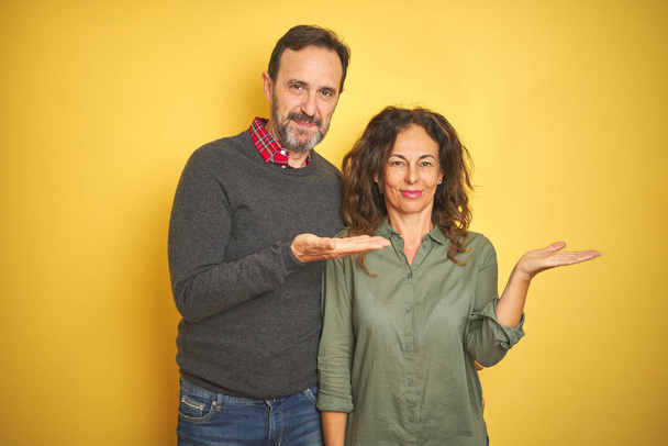 Beautiful middle age couple over isolated yellow background smiling cheerful presenting and pointing with palm of hand looking at the camera. - Фото, изображение