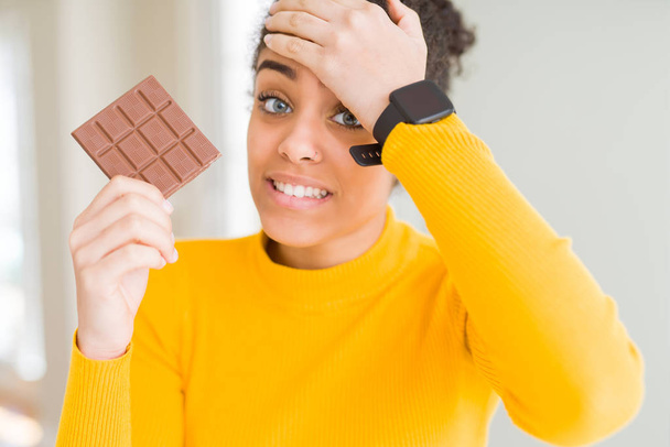 Молодая африканская американская девушка ест темный шоколад, как сладкая закуска, напряженная рукой на голове, шокированная стыдом и удивлением лица, сердитая и разочарованная. Страх и разочарование по ошибке
. - Фото, изображение