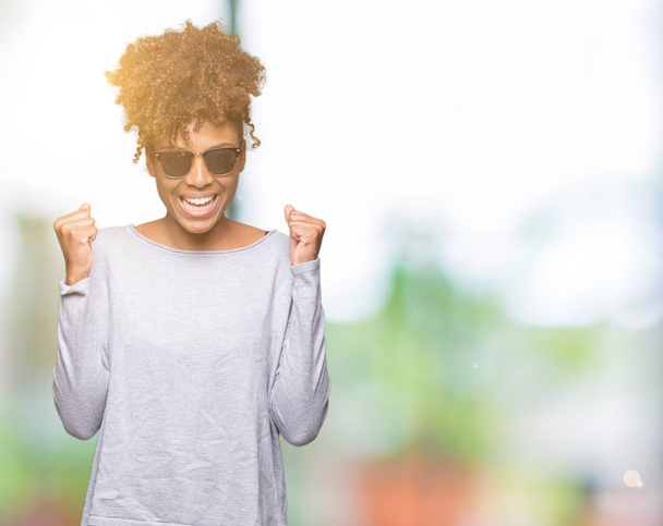Hermosa mujer afroamericana joven con gafas de sol sobre un fondo aislado celebrando sorprendida y sorprendida por el éxito con los brazos levantados y los ojos abiertos. Concepto ganador
. - Foto, imagen