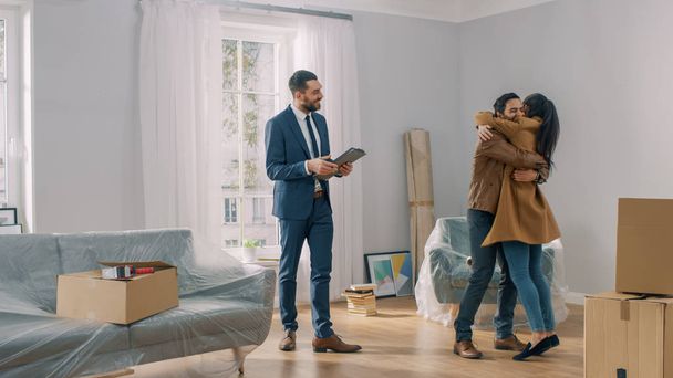 Real Estate Agent Shows Bright New Apartment to a Young Couple. Successful Young Couple Becoming Homeowners, Embraces and Hugs Each Other. Spacious Bright Home with Big Windows. - Photo, Image