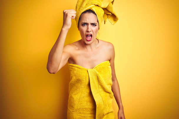 Belle femme portant une serviette de douche sur le corps et la tête sur fond jaune isolé en colère et fou levant le poing frustré et furieux tout en criant de colère. Rage et concept agressif
. - Photo, image
