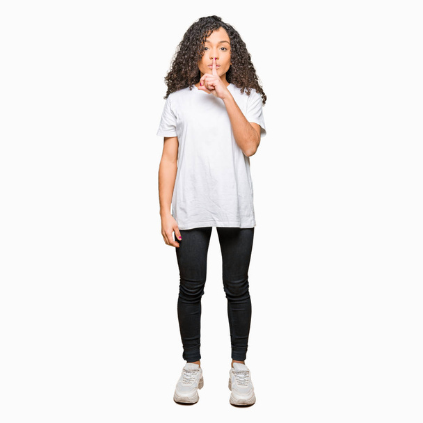 Young beautiful woman with curly hair wearing white t-shirt asking to be quiet with finger on lips. Silence and secret concept. - Photo, Image