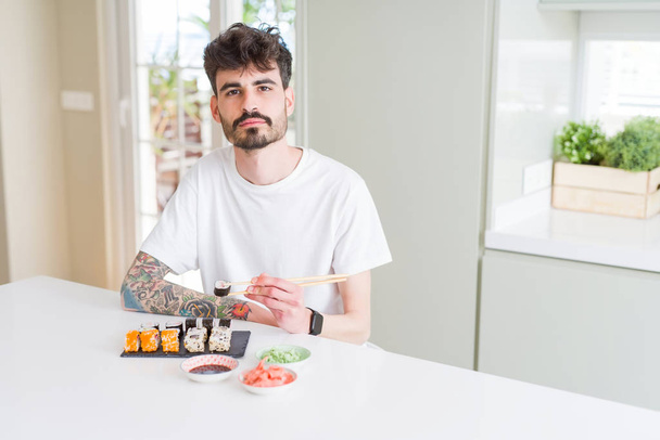 junger Mann isst asiatisches Sushi von der Lieferung nach Hause mit selbstbewusstem Gesichtsausdruck - Foto, Bild