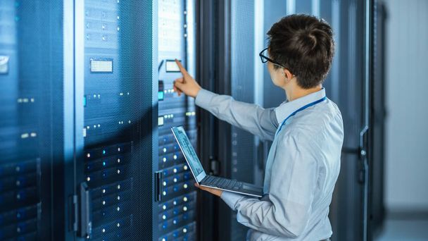 In the Modern Data Center: IT Engineer Standing Beside Open Server Rack Cabinets, Does Wireless Maintenance and Diagnostics Procedure with a Laptop. He Pushes Button. - Photo, Image