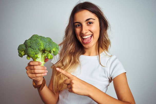 Young beautiful woman eating broccoli over grey isolated background very happy pointing with hand and finger - 写真・画像