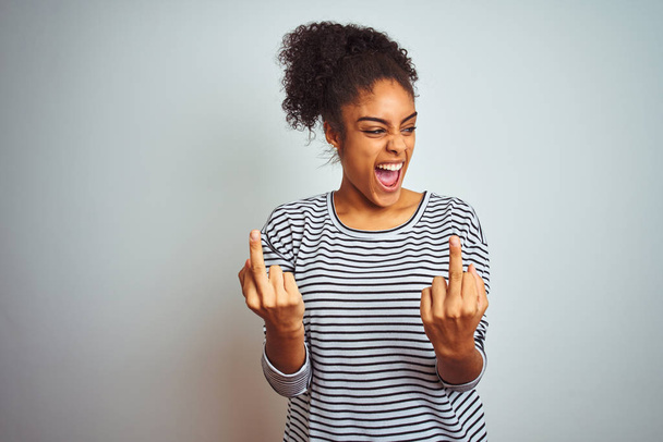 Mujer afroamericana vistiendo camiseta de rayas navy de pie sobre un fondo blanco aislado mostrando el dedo medio haciéndote follar mala expresión, provocación y actitud grosera. Gritando emocionado
 - Foto, Imagen