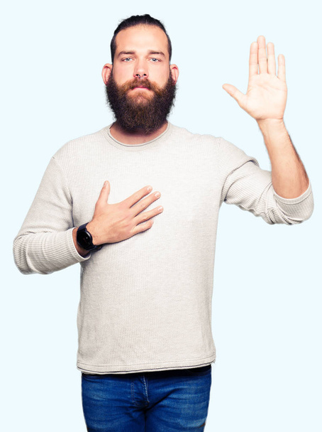Joven hombre rubio con suéter casual Juramento con la mano en el pecho y la palma abierta, haciendo una promesa de lealtad juramento
 - Foto, imagen