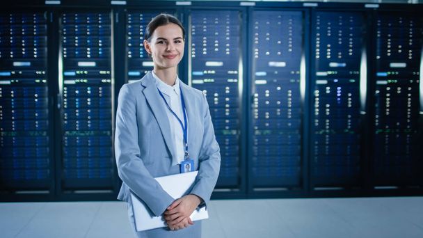 Female IT Specialist is Standing in Data Center Next to Server Racks, Holding Laptop and Smiling to the Camera. - 写真・画像