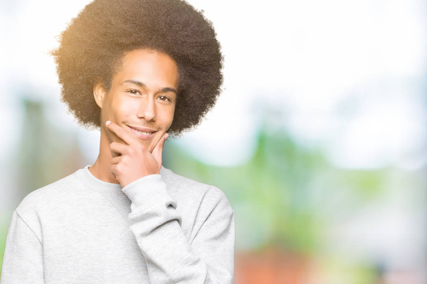 Giovane uomo afro americano con i capelli afro indossa felpa sportiva guardando fiducioso la fotocamera con il sorriso con le braccia incrociate e la mano alzata sul mento. Pensare positivo
. - Foto, immagini