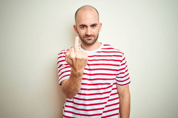 junger glatzköpfiger Mann mit Bart trägt lässig gestreiftes rotes T-Shirt über weißem, isoliertem Hintergrund und zeigt Mittelfinger, unhöflichen und unhöflichen Ausdruck - Foto, Bild