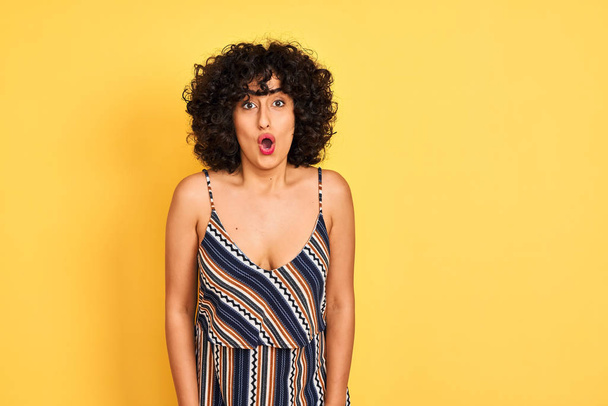 Arab woman with curly hair wearing striped colorful dress over isolated yellow background afraid and shocked with surprise expression, fear and excited face. - Photo, Image