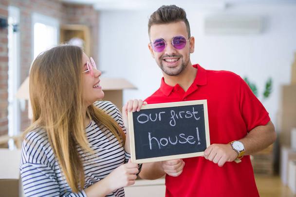 Νεαρό τρελό ζευγάρι φορώντας αστεία γυαλιά και κρατώντας πίνακα με το πρώτο μας κείμενο στο σπίτι, χαρούμενος και ενθουσιασμένος για τη μετακίνηση σε νέο διαμέρισμα - Φωτογραφία, εικόνα