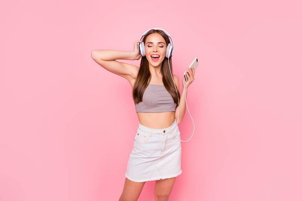 Portrait of lovely youth with her eyes closed holding modern technology wearing white skirt denim jeans grey tank-top isolated over pink background - Foto, Imagem