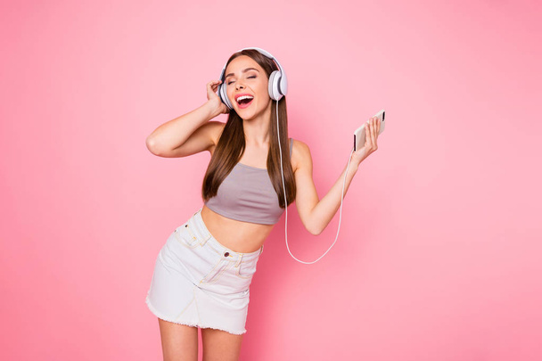Portrait of cute youth holding device closing eyes hearing playlist wearing trendy stylish tank-top denim jeans skirt isolated over pink background - 写真・画像