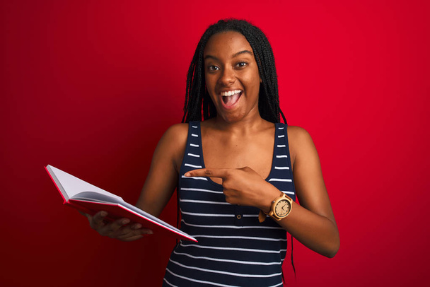 junge afrikanisch-amerikanische Studentin liest Buch steht über isolierten roten Hintergrund sehr glücklich zeigt mit Hand und Finger - Foto, Bild