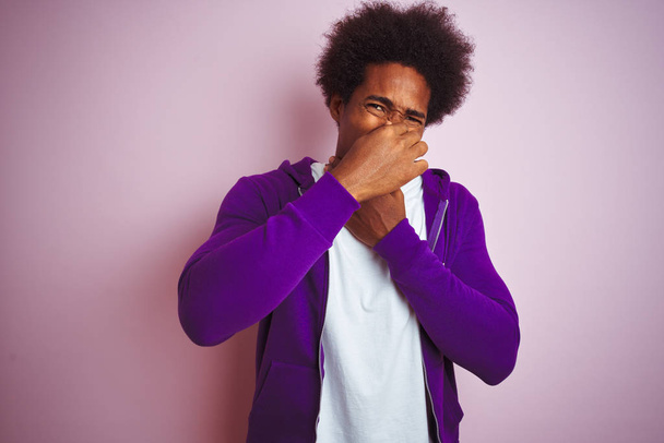 Młody African American Man ubrany fioletowy Bluza stoi na białym tle różowy zapach coś śmierdzące i obrzydliwe, nietolerancji zapach, trzymając oddech palcami na nos. Zła koncepcja zapachów. - Zdjęcie, obraz