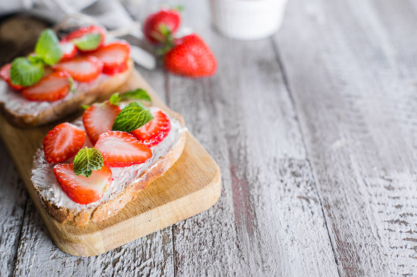 Two toasts or bruschetta with strawberry and mint on cream-cheese on wooden board on table background. Copy space - Fotografie, Obrázek