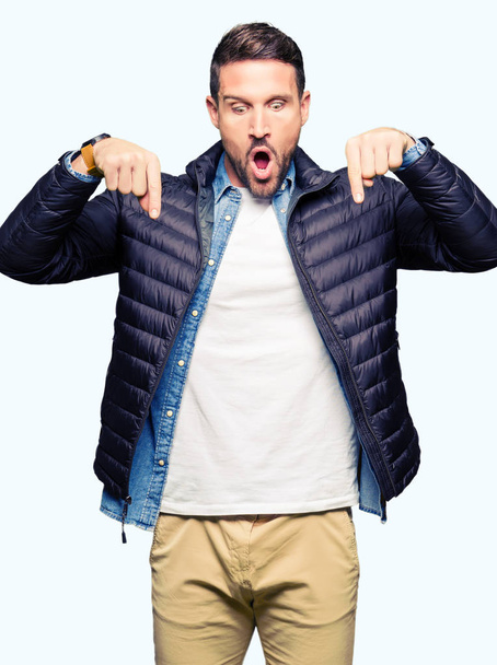 Pohledný muž nosit zimní kabát Pointing dolů s prsty ukazující reklama, překvapený obličej a otevřená ústa - Fotografie, Obrázek