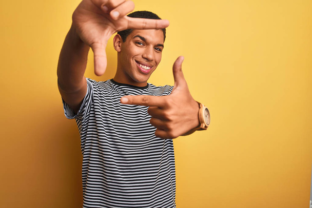 Hombre árabe guapo joven vistiendo camiseta de rayas navy sobre fondo amarillo aislado sonriendo haciendo marco con manos y dedos con la cara feliz. Concepto de creatividad y fotografía
. - Foto, imagen