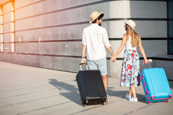 Two happy young tourists walking hand in hand away from the camera, pulling suitcases and looking at each other - Foto, Imagen
