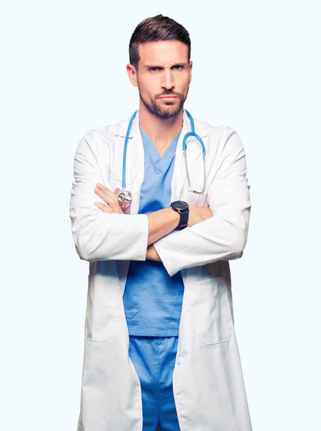 Przystojny lekarz człowiek ubrany medycznych uniform sceptyk na białym tle i nerwowy, dezaprobaty wyraz twarzy z skrzyżowanymi rękami. Negatywne osoba. - Zdjęcie, obraz