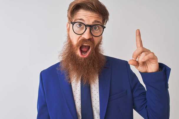Jonge Redhead Ierse zakenman dragen pak en bril over geïsoleerde witte achtergrond wijzende vinger omhoog met succesvolle idee. Verlaten en gelukkig. Nummer één. - Foto, afbeelding