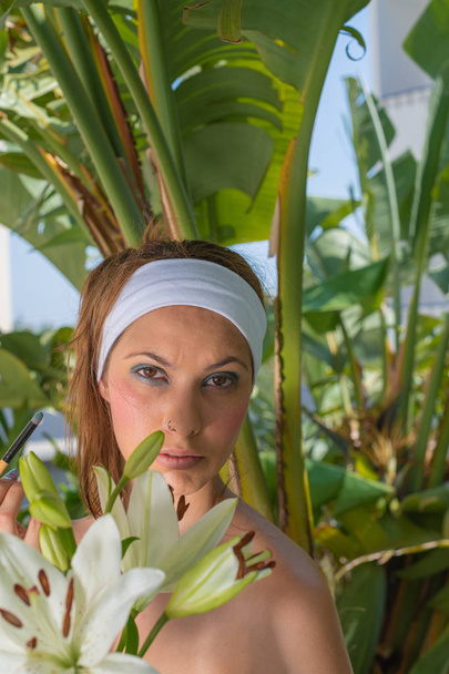 Woman posing with a green plant, showing her makeup in the garden - Young girl with a white scarf on her head - Foto, afbeelding