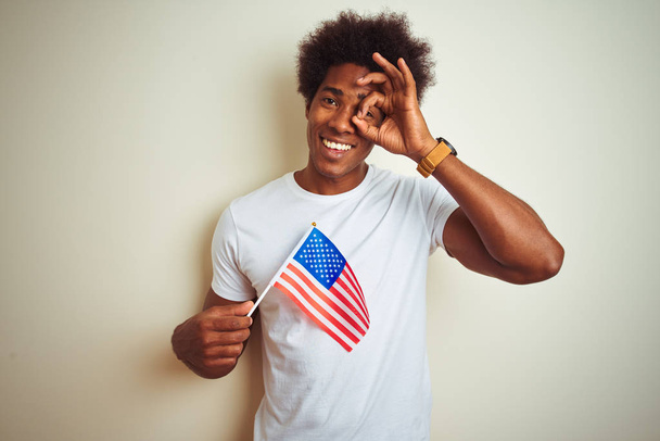 Афро людина Холдинг Сполучені маєтності Америки Прапор США стояти над ізольованим білим фоном з щасливою особою посміхаючись робити OK підписати з рукою на око дивиться через пальці - Фото, зображення
