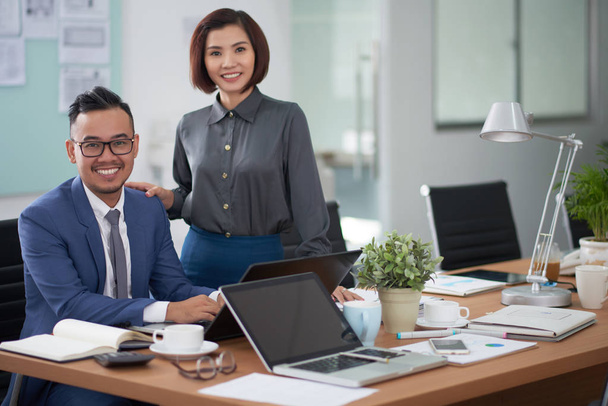 Portret azjatyckich młody biznesmen siedzi na biurku wpisując na laptopie z piękną bizneswoman stoi w pobliżu przez niego, uśmiechają się do kamery w biurze - Zdjęcie, obraz