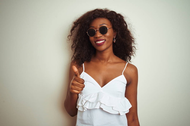 Giovane donna afroamericana che indossa t-shirt e occhiali da sole su sfondo bianco isolato facendo felice pollice in su gesto con la mano. Approvazione espressione guardando la fotocamera con successo mostrando
. - Foto, immagini