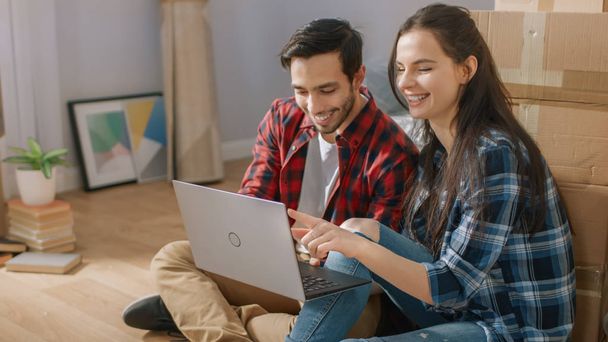 Happy Young Couple Sitting on the Floor of the Newly Rented / Purchased Apartment Use Laptop Computer. Unpacked Cardboard Boxes and Covered Furniture in the Modern Bright Sweet Home. - 写真・画像