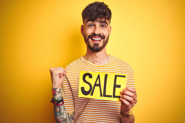 Young man with tattoo holding sale banner standing over isolated yellow background screaming proud and celebrating victory and success very excited, cheering emotion - Zdjęcie, obraz