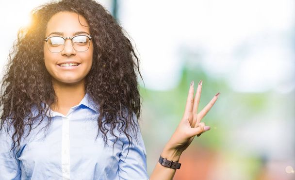 Nuori kaunis liiketoiminnan tyttö kihara tukka yllään lasit osoittavat ja osoittaa sormilla numero kolme hymyillen itsevarma ja onnellinen
. - Valokuva, kuva