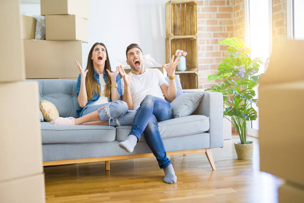 Young beautiful couple relaxing sitting on the sofa around boxes from moving to new house crazy and mad shouting and yelling with aggressive expression and arms raised. Frustration concept. - Foto, Bild