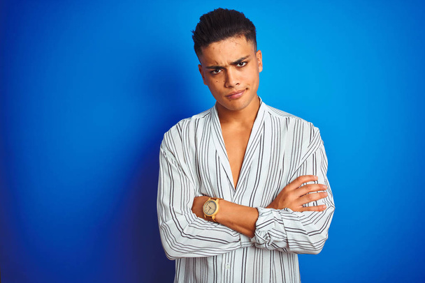Young brazilian man wearing striped shirt standing over isolated blue background skeptic and nervous, disapproving expression on face with crossed arms. Negative person. - Photo, Image