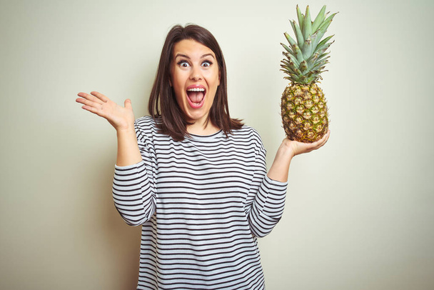 Young beautiful woman holding tropical fruit pineapple over isolated background very happy and excited, winner expression celebrating victory screaming with big smile and raised hands - Zdjęcie, obraz